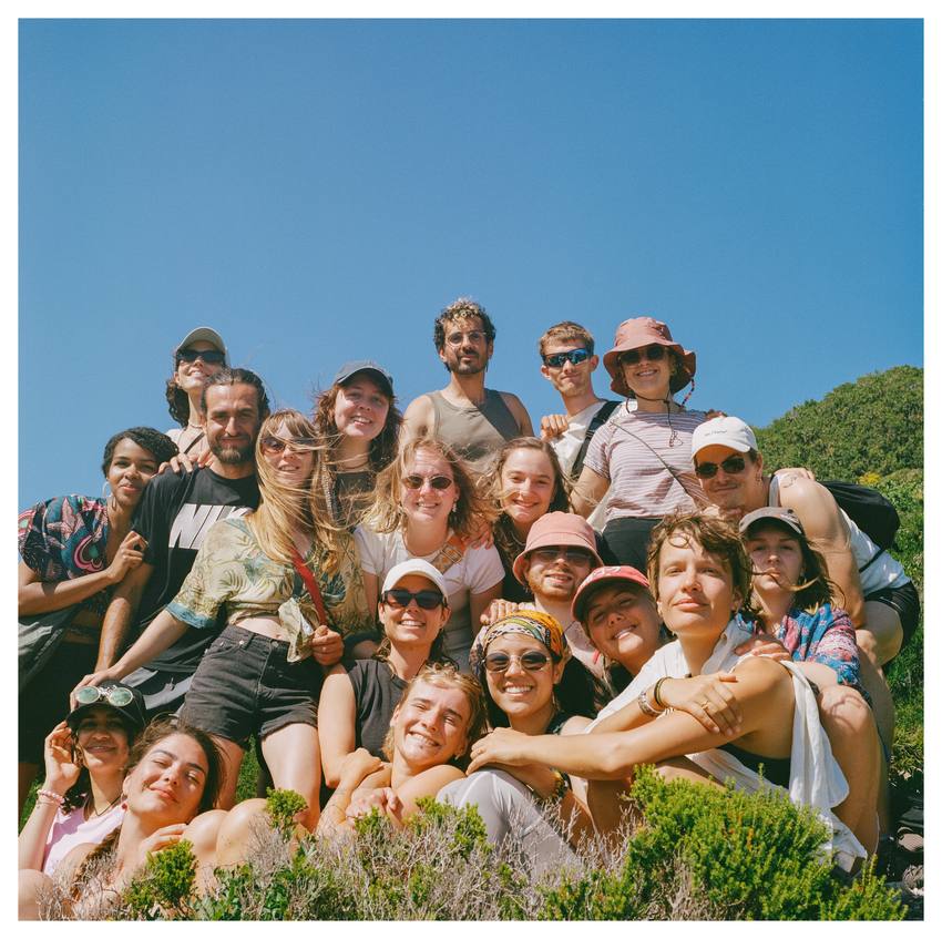 A big group of friends piled together on a sunny hillside, laughing at the camera