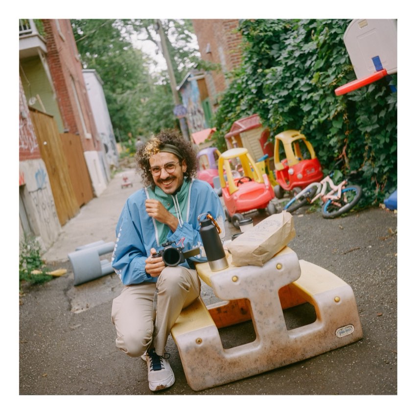 Karam crouching in a colourful alley in his blue jacket, camera in hand, smiling at the lens
